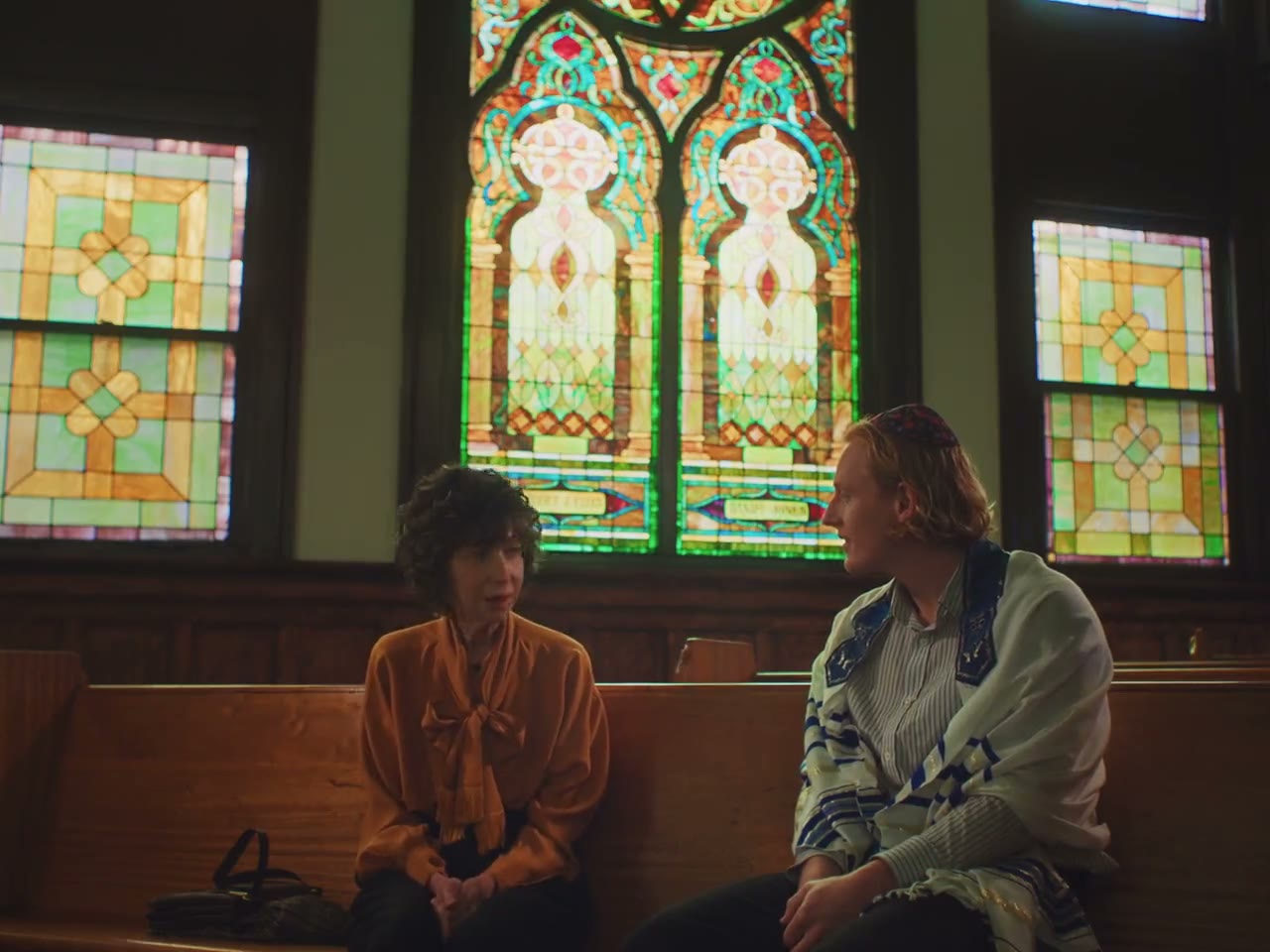 Frame from Beatrix Is Invisible — two people seated on a wooden pew in front of tall stained-glass windows